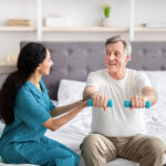 Young rehabilitation therapist assisting senior male patient with strength exercises with dumbbells on bed at home